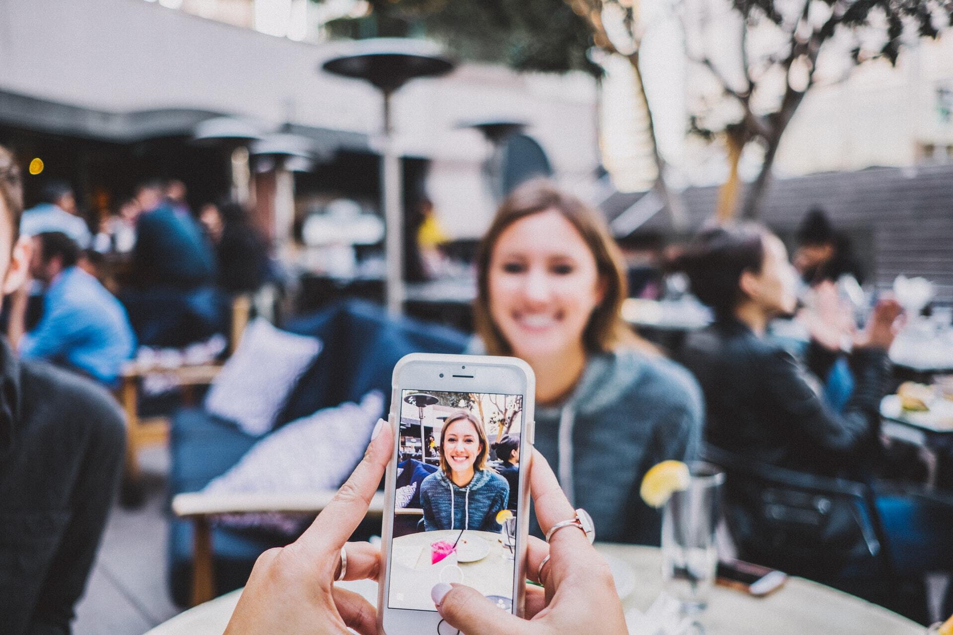 Photo of a photo being taken of a woman on a smart phone at an outdoor cafe